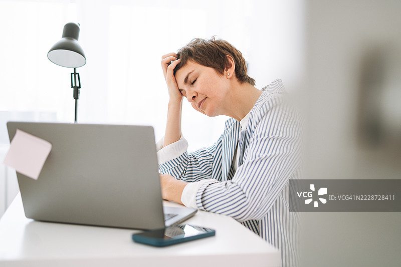 疲惫的女商人在办公室加班，虽然疲惫但专注于笔记本电脑，体现了职场疲劳的概念图片素材