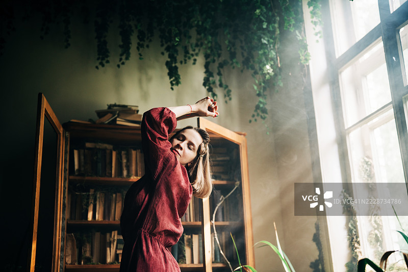 一位身着深红色连衣裙的年轻女子，站在阳光明媚的房间里，四周是旧书和茂密的植物，若有所思。图片素材