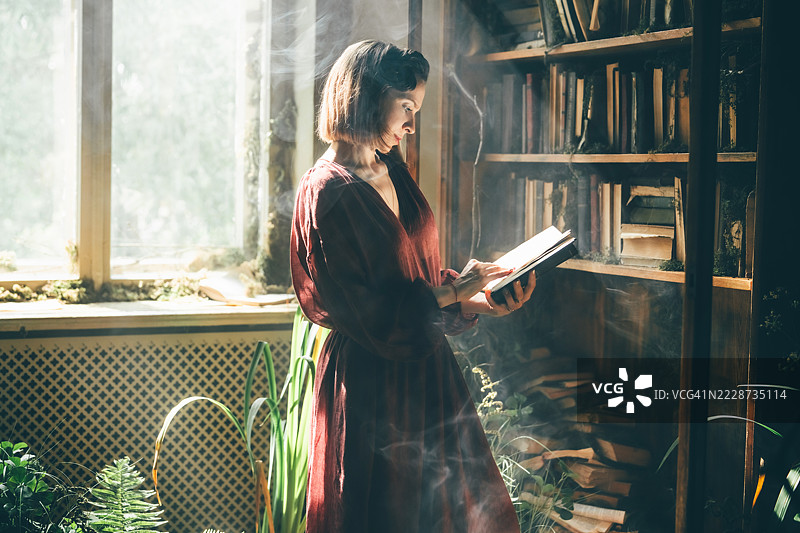 一位穿着深红色裙子的女性在阳光明媚的房间里沉思，房间里满是古老的书籍和郁郁葱葱的植物。图片素材