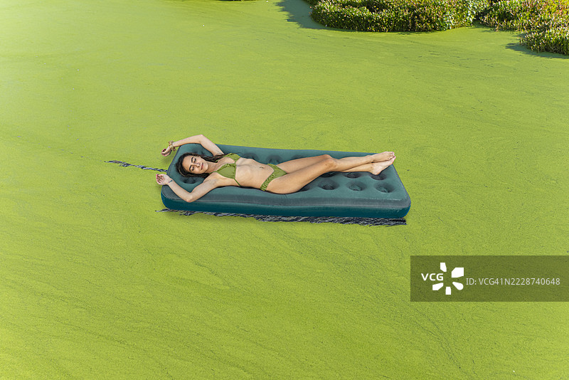 女人在一个小湖中漂浮的充气床垫上，湖面覆盖着绿色藻类。图片素材