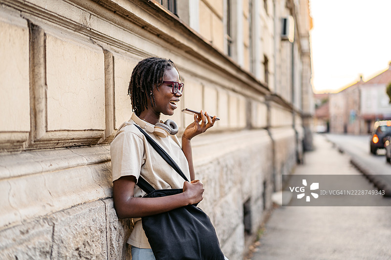 年轻的黑人女性在城市街道上靠着建筑物用手机发送语音信息图片素材