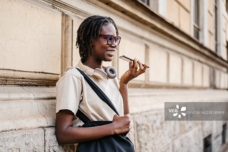 年轻的黑人女性在城市街道上靠着建筑物，用手机发送语音信息图片素材