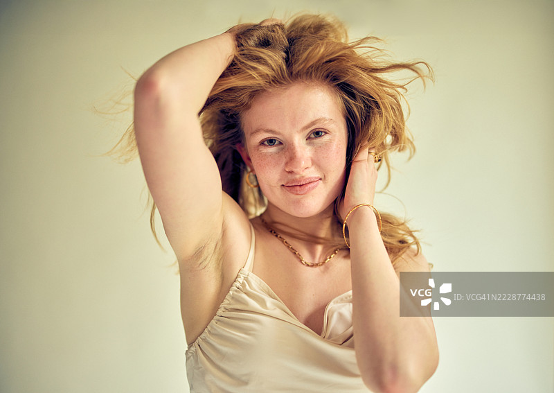 自信的年轻红发女性在无袖上衣中摆姿势，双手插在头发里，面对镜头拍摄的工作室肖像。图片素材