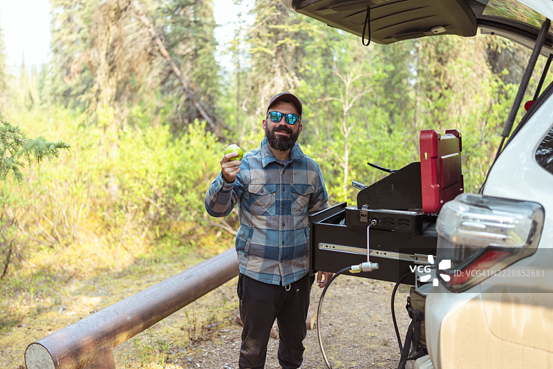 一个微笑的男人在露营时站在他的SUV旁边，直视镜头。图片素材