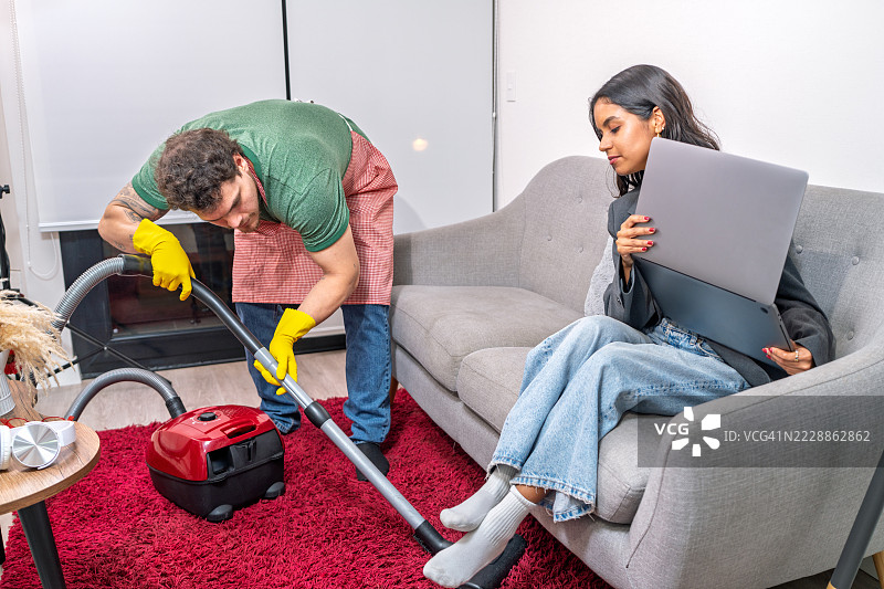 男人在家里吸尘，女人在笔记本电脑上工作图片素材
