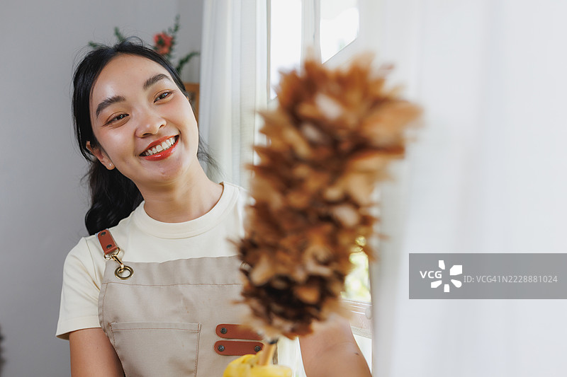 年轻的亚洲女性用羽毛掸子清洁白色窗帘，面带微笑看向镜头图片素材