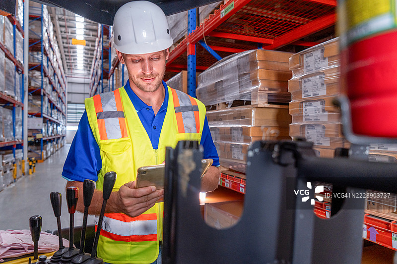 零售仓库内满是货架和商品：一名男子驾驶电动叉车准备将装有纸箱的托盘提升到货架上。人们在配送中心工作，使用叉车。图片素材
