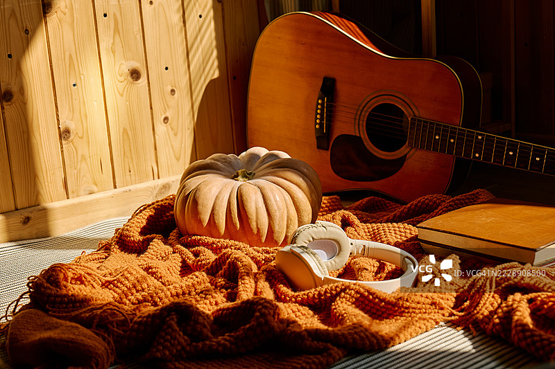 阳光下的吉他、书籍和南瓜，放在橙色毛呢格子布上，背景是木墙。秋季氛围概念图片素材