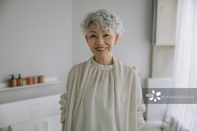 微笑的日本老年女性站在明亮的极简风格家居中，注视着镜头图片素材