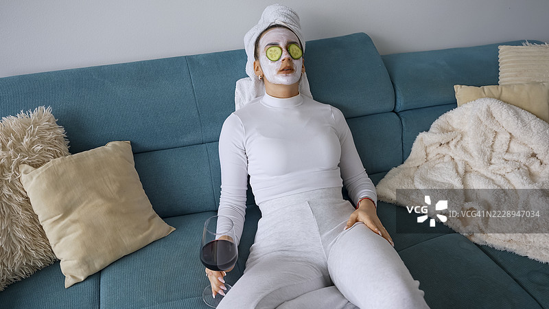 年轻女性在沙发上放松，面带面膜，手持一杯红酒图片素材