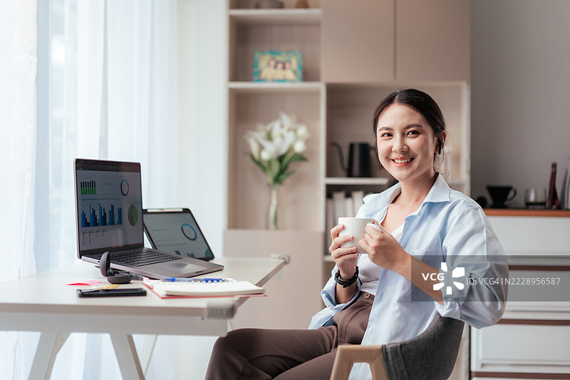 愉快的亚洲女性在家工作时微笑，手握咖啡杯，坐在舒适的桌子前，旁边有笔记本电脑和平板电脑。适合远程工作的家庭办公室布置，数字生活方式与工作生活平衡。图片素材