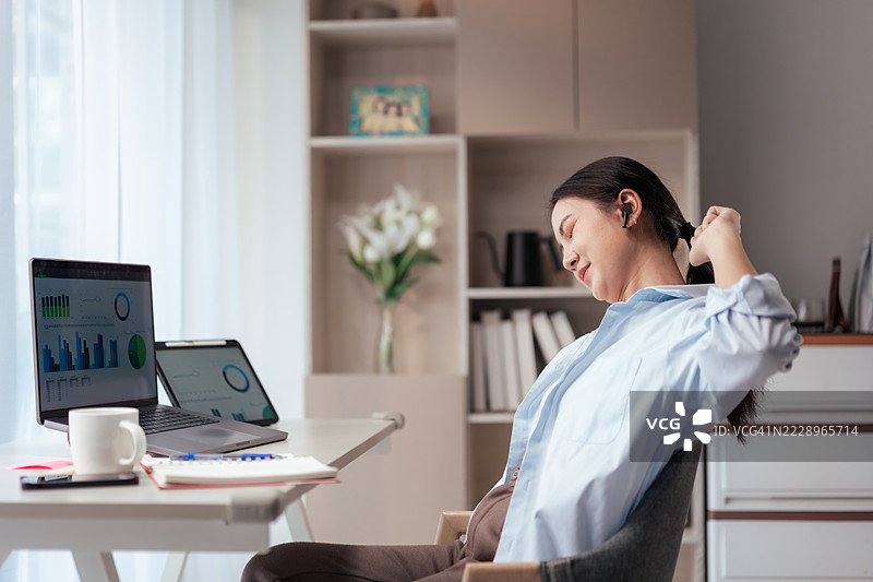 亚洲女性在家中办公室休息时伸展和放松。轻松的远程工作生活方式，配有数字设备、笔记本电脑、笔记本和咖啡杯。舒适的自由职业工作空间。图片素材