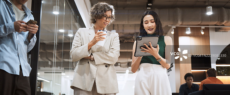 高管与亚洲女性在城市办公室的走廊里交谈，手持数字平板，旁边有同事。全球商业、市场营销、金融、科技、多元文化的现代概念。图片素材