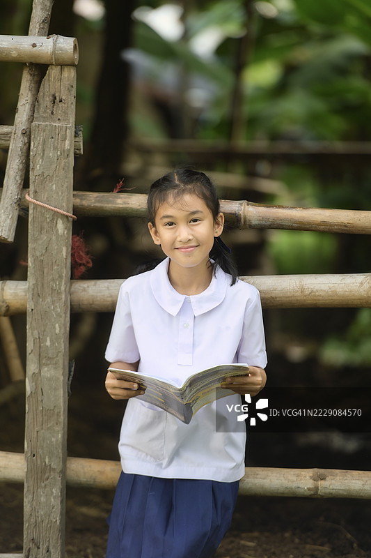 微笑的女孩穿着校服，坐在木栅栏上阅读一本书，泰国图片素材
