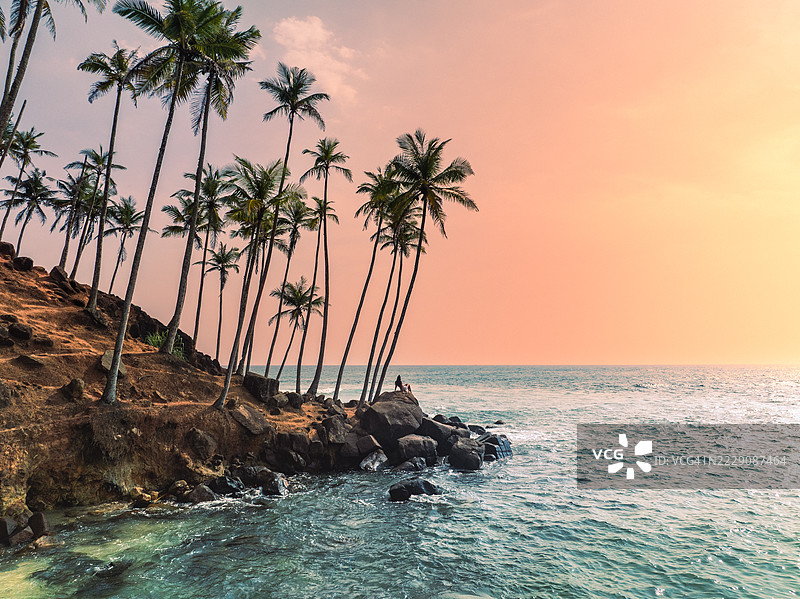 航拍图像显示一位女性坐在岩石上，夕阳下眺望印度洋，地点为斯里兰卡南部省马塔拉区米里萨的椰子树山。图片素材