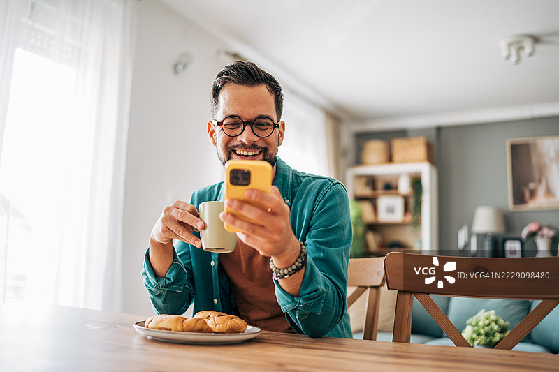 快乐的男人在家早餐时使用智能手机并喝咖啡图片素材