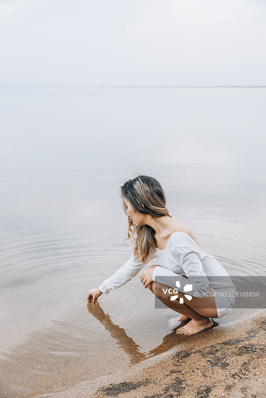 一个无法辨认的女人赤脚蹲在湖边，用手指拨动水面，凝视着水面。图片素材