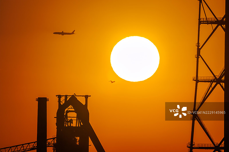 第一缕阳光与一架客机和鸟的剪影。它即将降落在附近的羽田机场。第一缕阳光从运河对岸的烟囱和电厂、钢铁厂上升。横滨，神奈川。图片素材