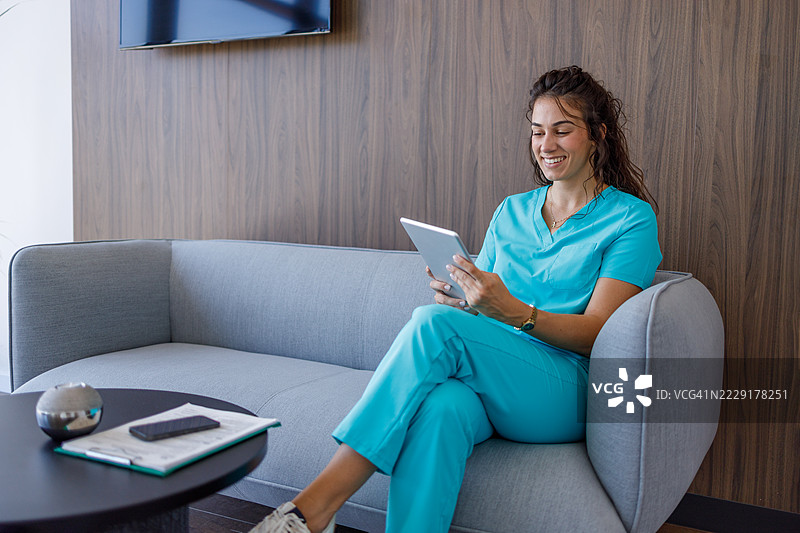 微笑的护士坐在医院候诊室的沙发上使用平板电脑图片素材