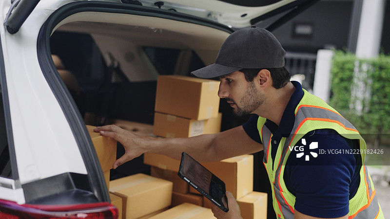快递员穿着蓝色 polo 衫，正在查看数字平板电脑以获取纸箱包裹的追踪信息，涉及快递物流和快递服务流程。图片素材
