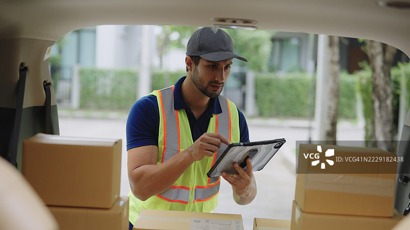 穿着蓝色 polo 衫的快递员正在查看数字平板电脑，以获取纸箱包裹的追踪信息，涉及快递物流和快递服务流程。图片素材
