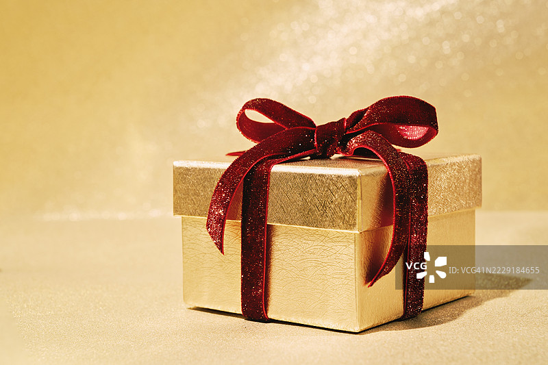 奢华的金色礼品盒，上面系着红色天鹅绒丝带蝴蝶结，背景为金色闪光。侧面视图图片素材