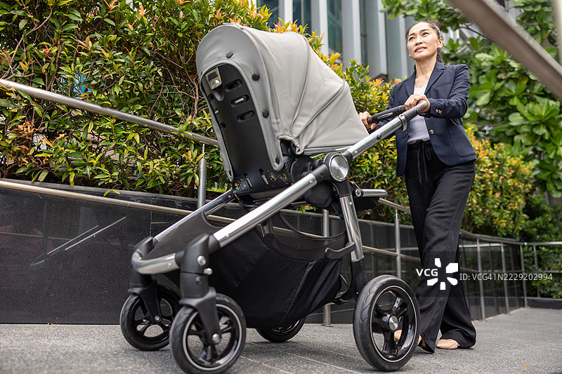 微笑的亚洲女性穿着商务服装，在户外的人行道上推着婴儿推车，周围是郁郁葱葱的植物，展现了在城市环境中平衡母亲角色与职业生涯的状态。图片素材