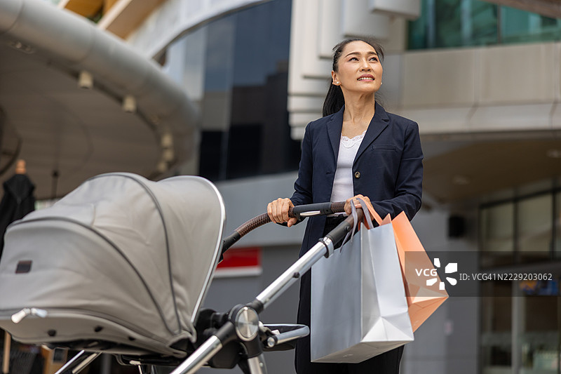 微笑的亚洲女性穿着商务服装，一边推着婴儿车，一边提着多个购物袋，站在现代购物中心外，展现了城市生活方式和多任务的母亲角色。图片素材