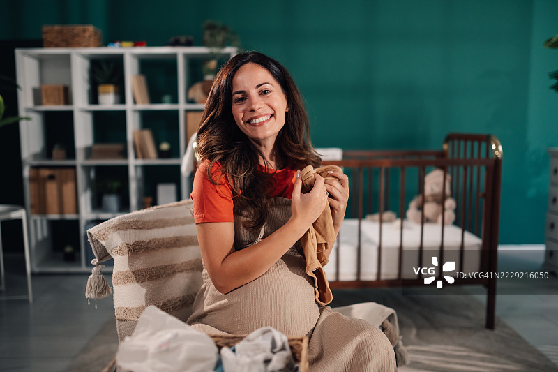 怀孕的女人在家微笑着拿着婴儿衣物图片素材
