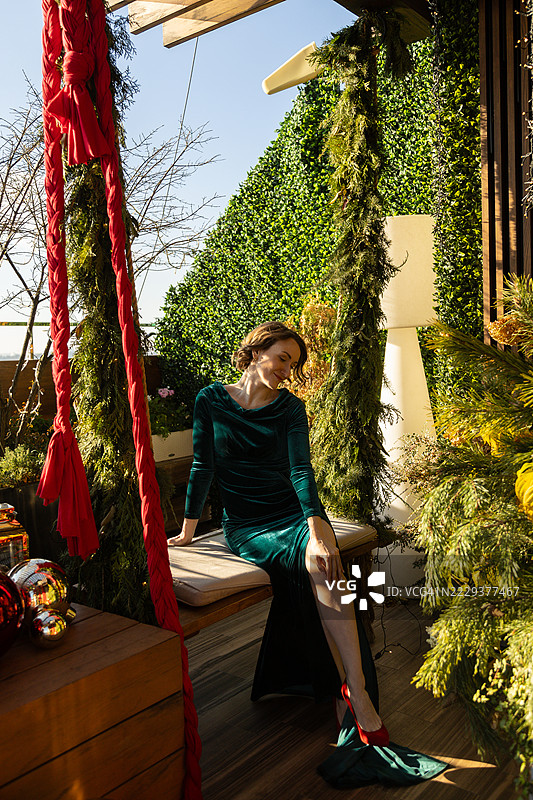 微笑的女性穿着开衩长裙，坐在节日气氛浓厚的屋顶秋千椅上。图片素材