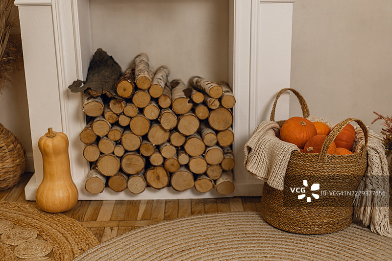 白色壁炉内堆叠的桦木柴，旁边有一个装着橙色南瓜的篮子和一条编织毛毯。图片素材
