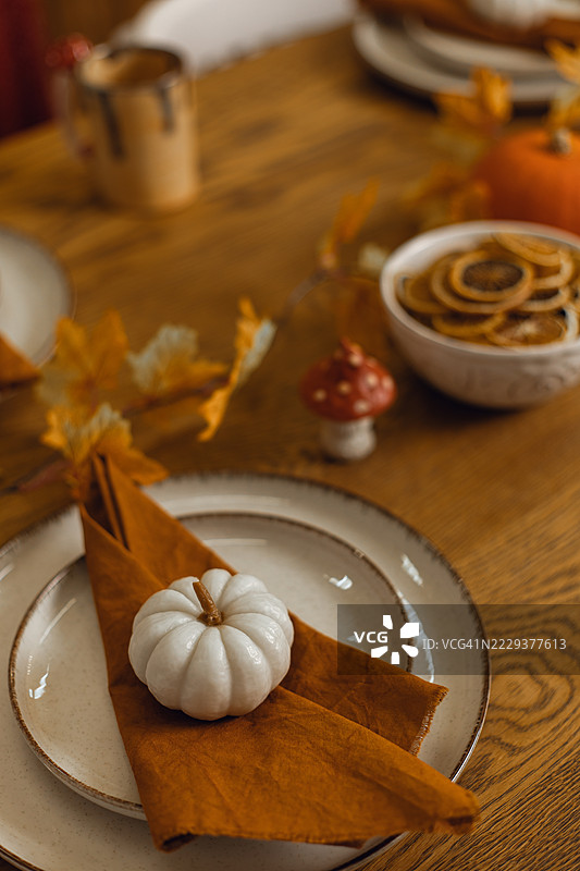 秋季餐桌上放着一只白色迷你南瓜，置于折叠的餐巾上，周围环绕着秋天的落叶和一个装饰性蘑菇，还有一碗干橙片。图片素材