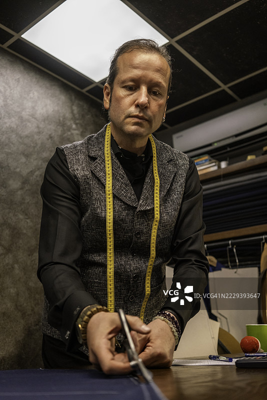 男性时装设计师正在裁剪面料，低角度拍摄，测量工具挂在他的脖子上，展现小型企业的静态场景。图片素材