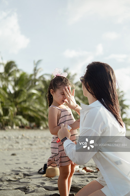 母亲在女儿的脸上涂抹防晒霜，保护孩子的皮肤在阳光明媚的海滩度假期间。图片素材