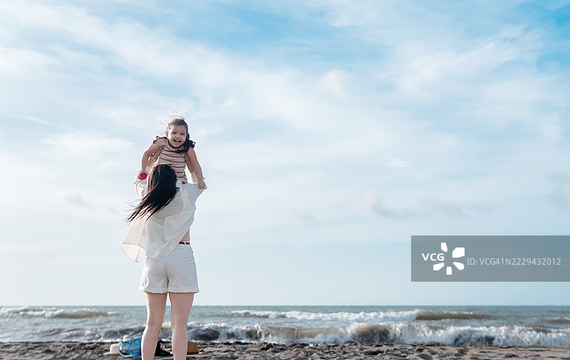 母亲在阳光明媚的海滩上顽皮地将女儿举起，创造了一个温馨的家庭亲密时刻，尽享他们的夏季假期。图片素材