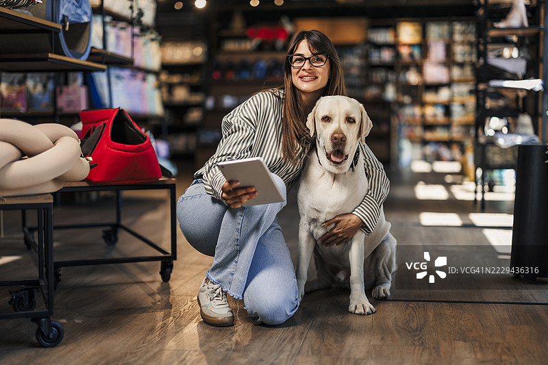 女员工在宠物店使用平板电脑，旁边有一只狗在走动。图片素材