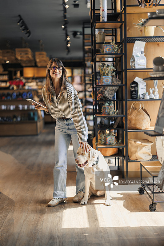 女员工在宠物店工作时使用数字平板电脑，旁边有一只狗在走动。图片素材