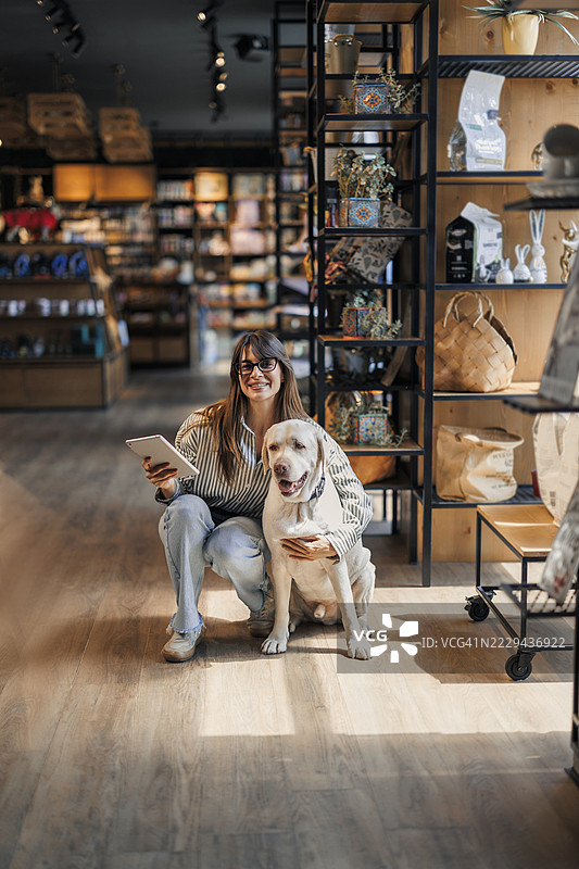 女员工在宠物店工作时使用数字平板电脑，旁边有一只狗在走动。图片素材