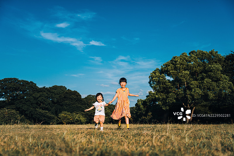 两个快乐的亚洲姐妹手牵手在草地上奔跑，享受大自然。女孩们在一个美好的日子里在公园里玩得很开心。兄妹之间的爱与团结。家庭的爱与关怀。在大自然中探索和户外乐趣。图片素材