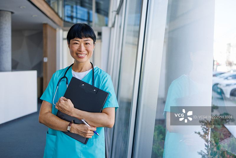 微笑的亚洲医生穿着手术服，在医院走廊里拿着夹板。图片素材