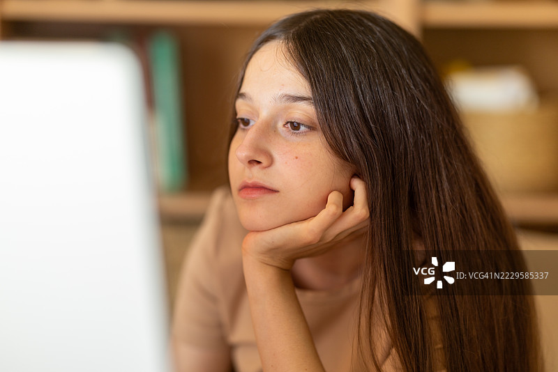 年轻女学生倚靠在桌子上，沉思地凝视着笔记本电脑屏幕，面露沉思的表情图片素材