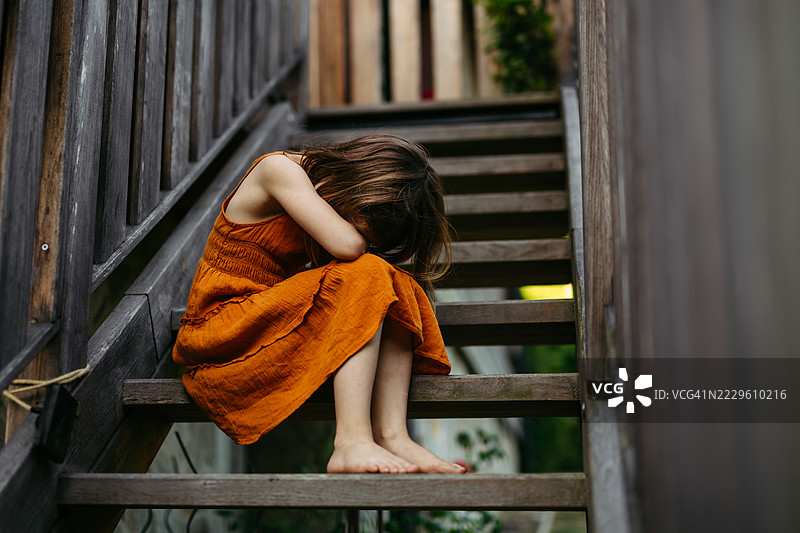 穿着橙色裙子的女孩坐在木楼梯上哭泣。图片素材
