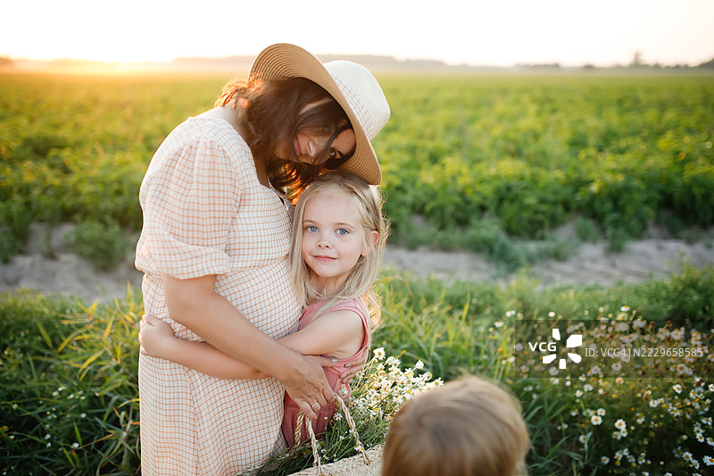 关爱时刻：怀孕的母亲与她的长发金发女儿在日落时分相拥，周围充满了对新奇迹的期待。两人面向镜头。图片素材