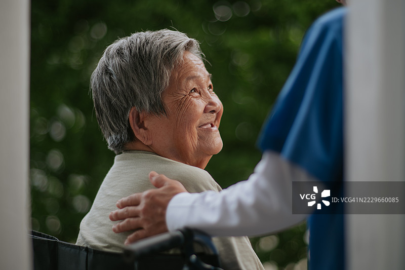 透过窗帘安慰坐在轮椅上的老年女性的护理人员。图片素材