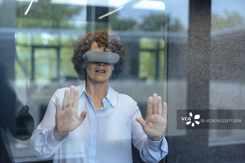 通过玻璃看到的商务女性佩戴虚拟现实模拟器并做手势图片素材