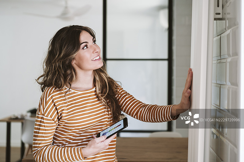 年轻女性在家中使用智能手机应用并触摸暖气片图片素材