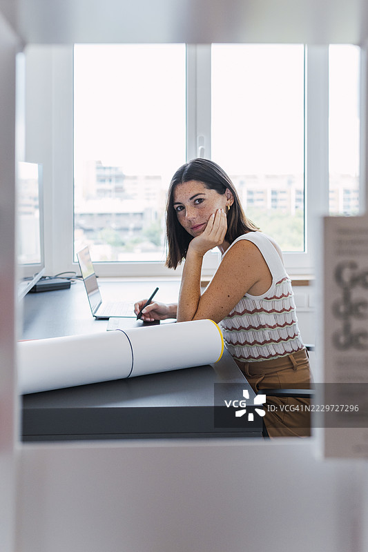 微笑的年轻女商人坐在桌面前，手托着下巴。图片素材