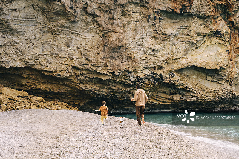 年轻父亲、小男孩和狗在沙滩上奔跑，背景是美丽的岩石纹理。图片素材