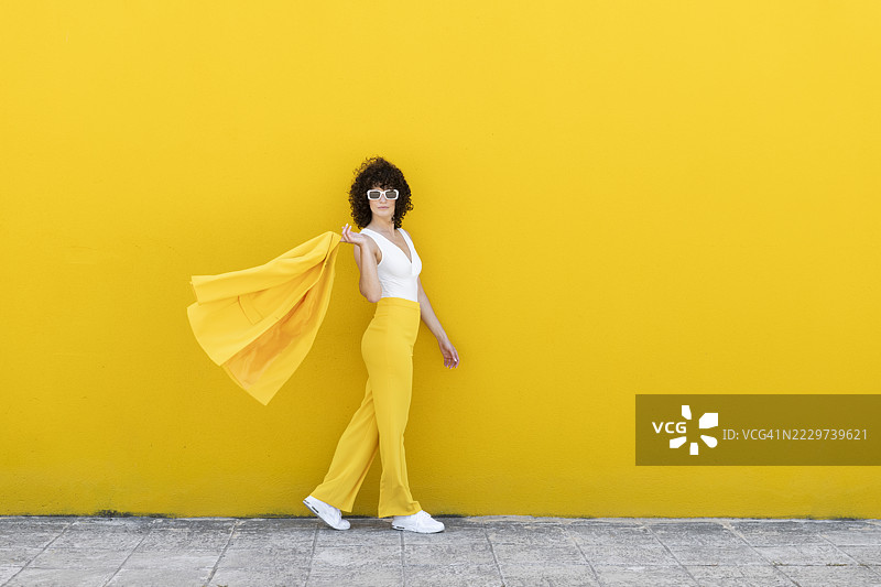 时尚女性穿着西装外套，戴着太阳镜，走在人行道上，背景是黄色墙壁。图片素材