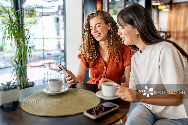 两位年轻女性在咖啡店里欢笑并使用智能手机图片素材